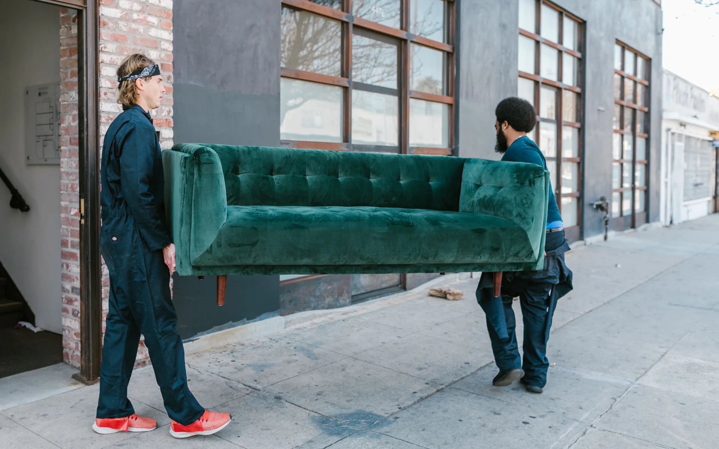 Two movers carrying a large tufted sofa together along a sidewalk